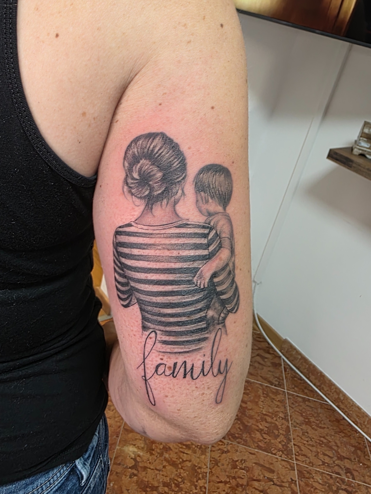 Familienmotiv Tattoo – Mutter mit Kind im Arm, Schriftzug ‚Family‘ darunter, auf Trizeps – Saints Tattoo Pirna